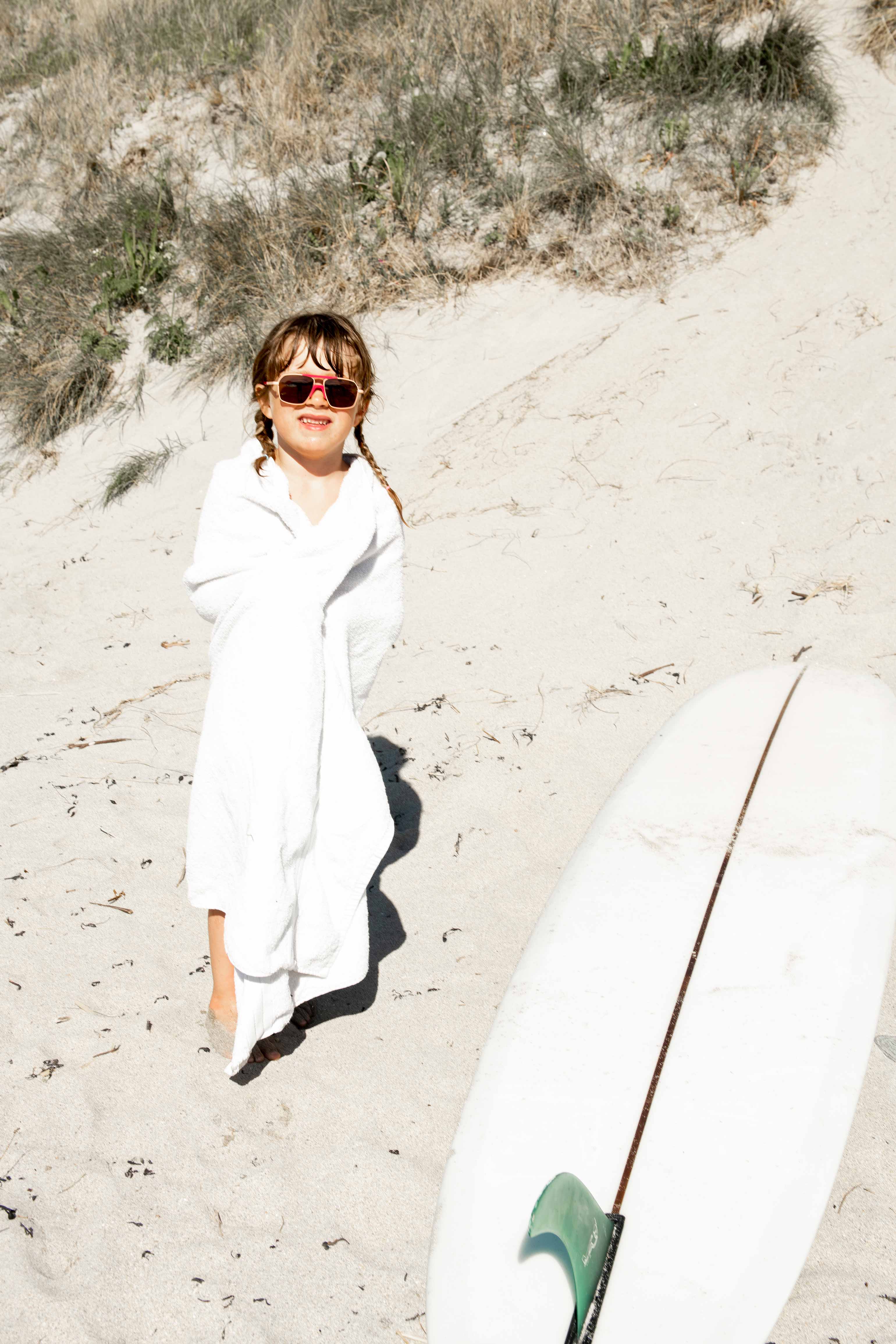 Young girl in a white towel standing on a sandy beach with a surfboard nearby wearing Mokki sunglasses for kids