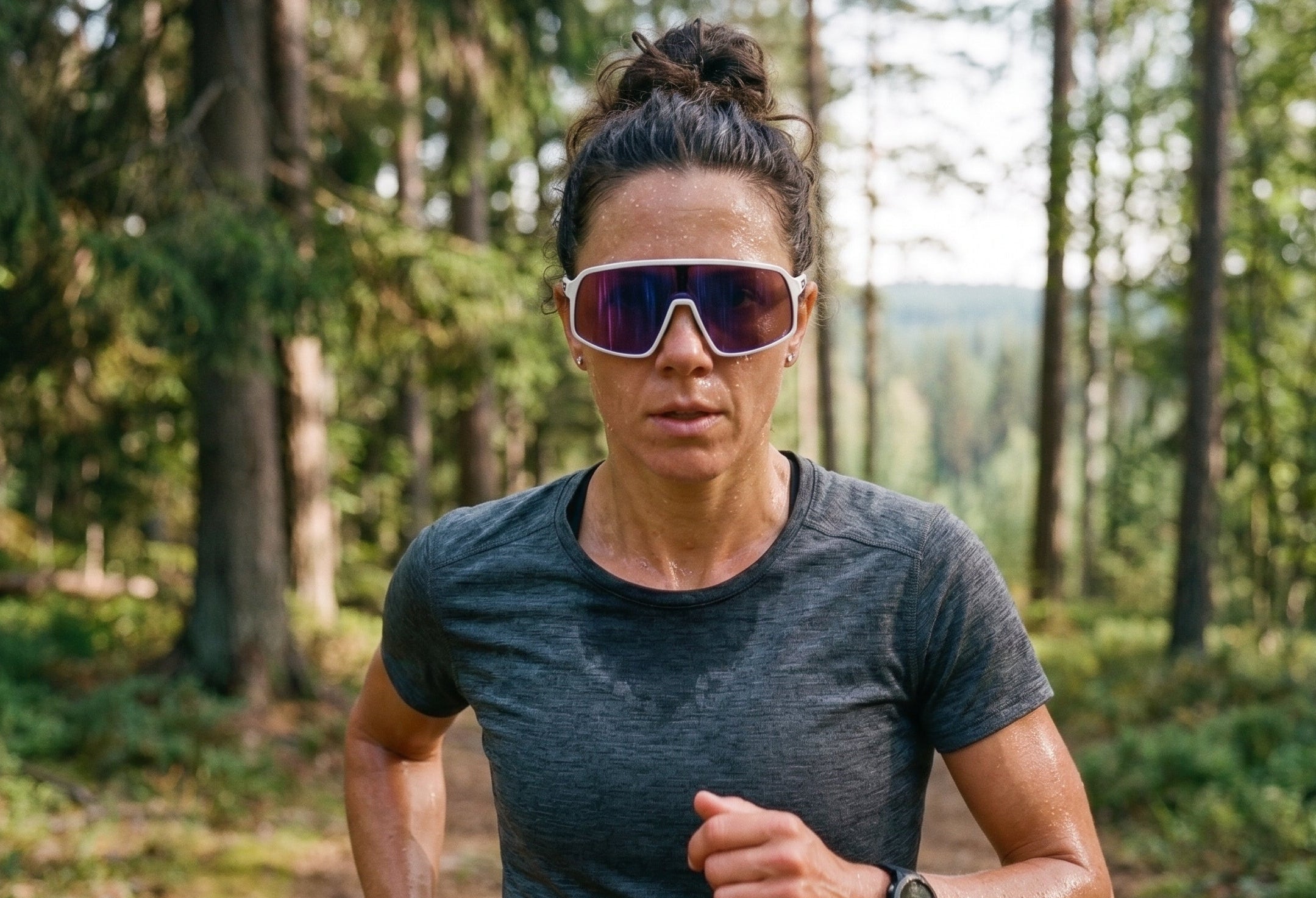 A woman running through a forest wearing oversized shield sunglasses with a white frame and mirrored lenses, a dark gray athletic t-shirt, and a smartwatch.