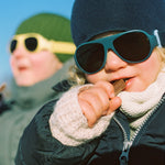 Child wearing Click & Change sunglasses and winter clothing against a blue sky eating Kvikk Lunsj chocolate 