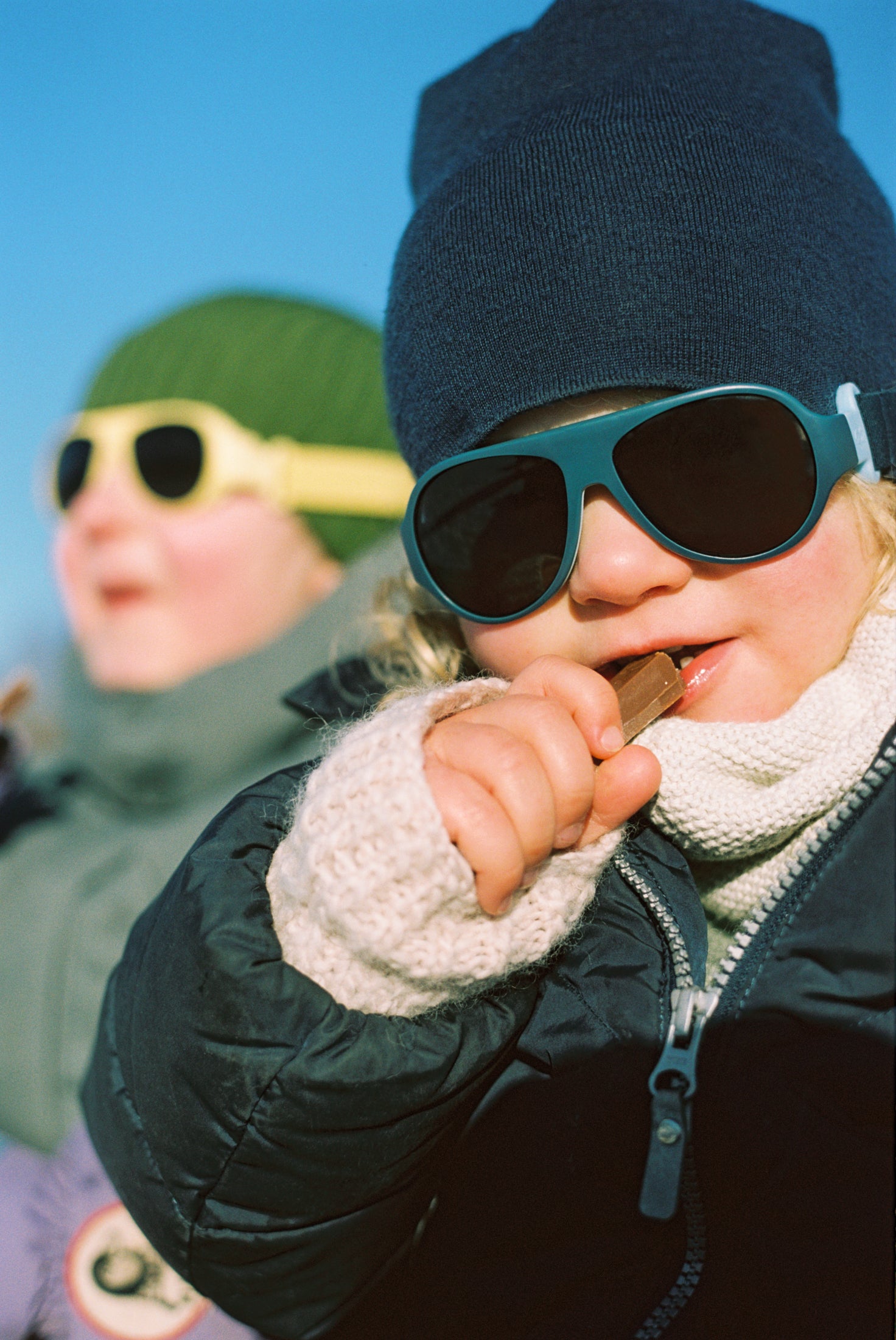 Child wearing Click & Change sunglasses and winter clothing against a blue sky eating Kvikk Lunsj chocolate 