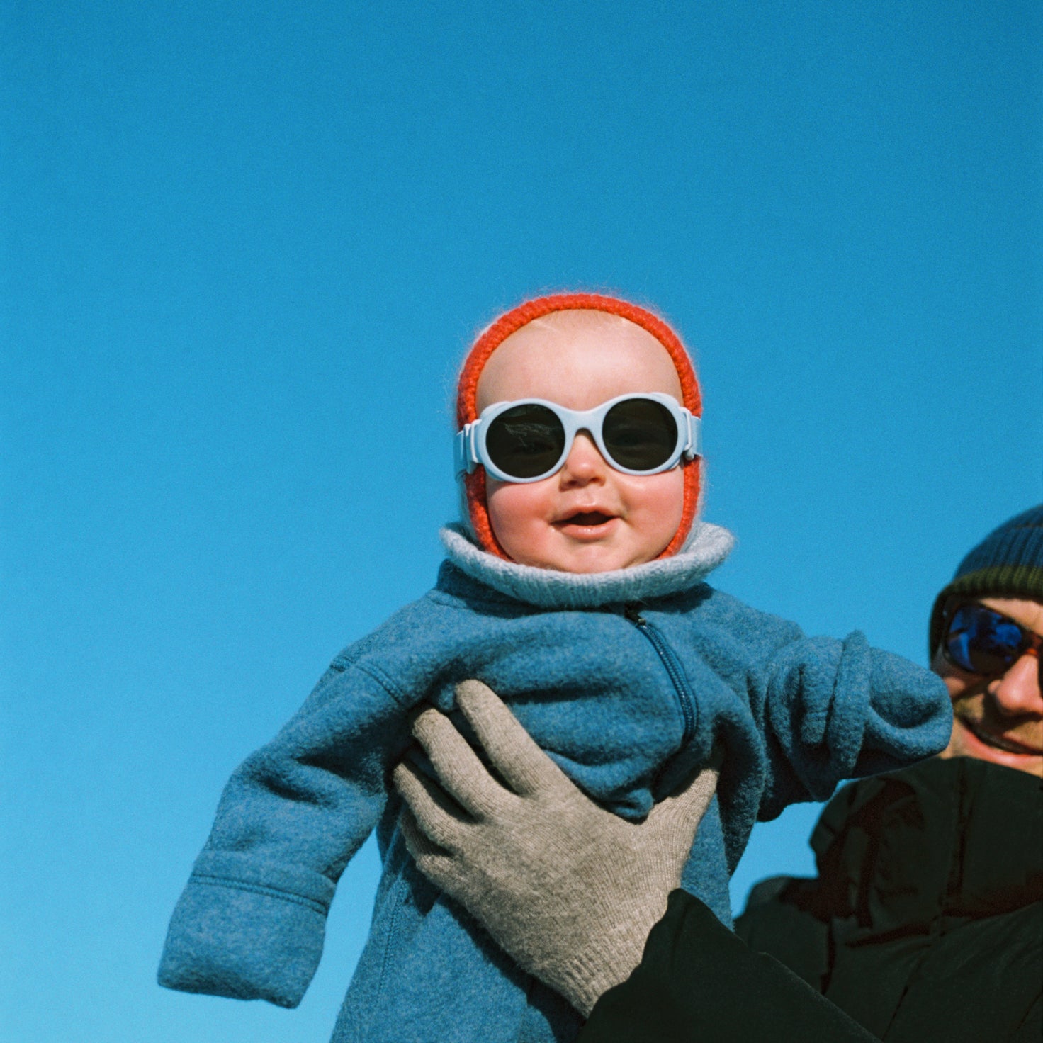 Baby in blue coat and red hat with Mokki Click & Change sunglasses held by an adult against a clear blue sky.