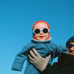 Baby in blue coat and red hat with Mokki Click & Change sunglasses held by an adult against a clear blue sky.
