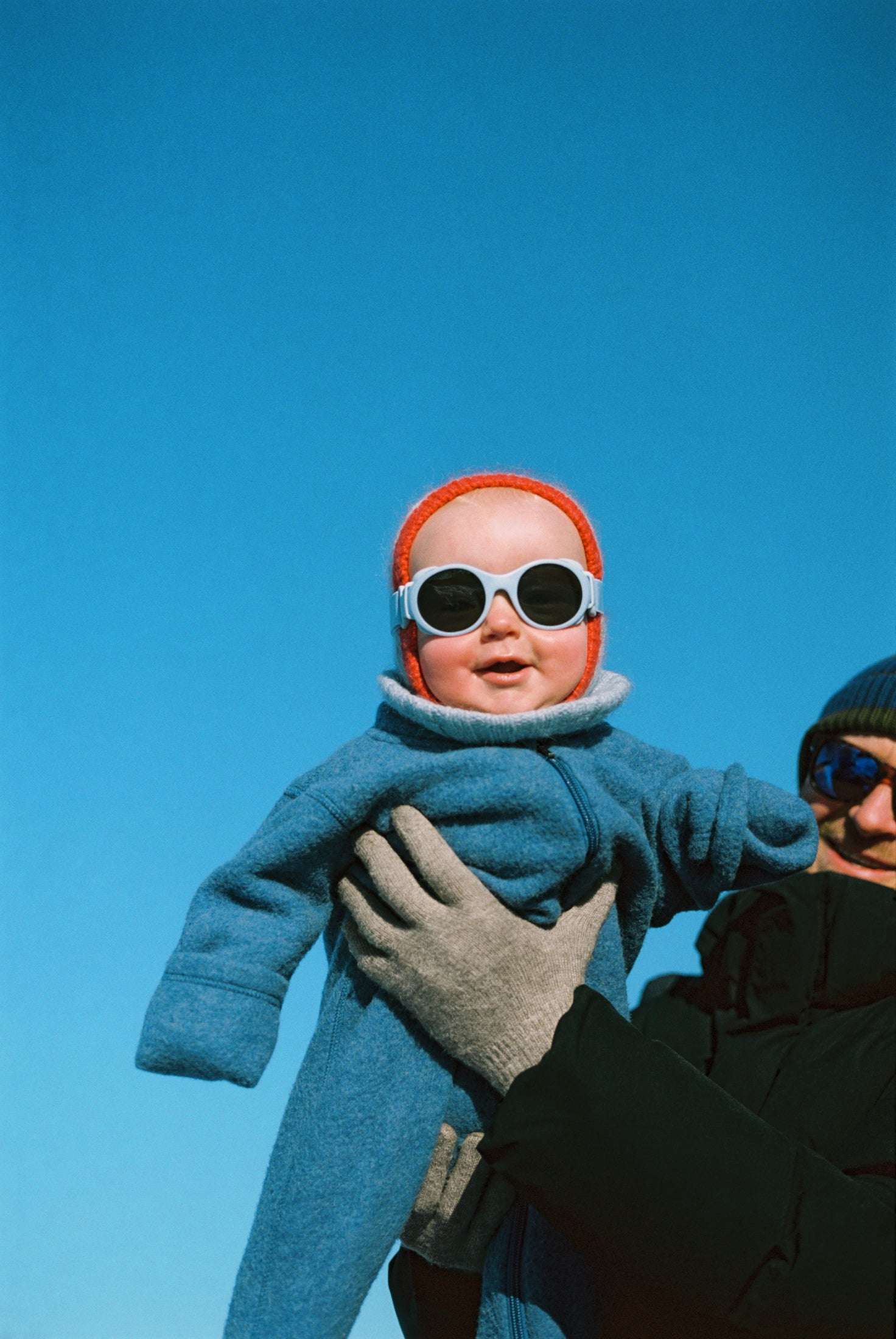 Baby in blue coat and red hat with Mokki Click & Change sunglasses held by an adult against a clear blue sky.