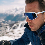 A man in a snowy mountain setting wearing white-framed ski sunglasses with blue mirrored lenses, a blue jacket, and black gloves. Film border graphics are visible on the left and right edges.