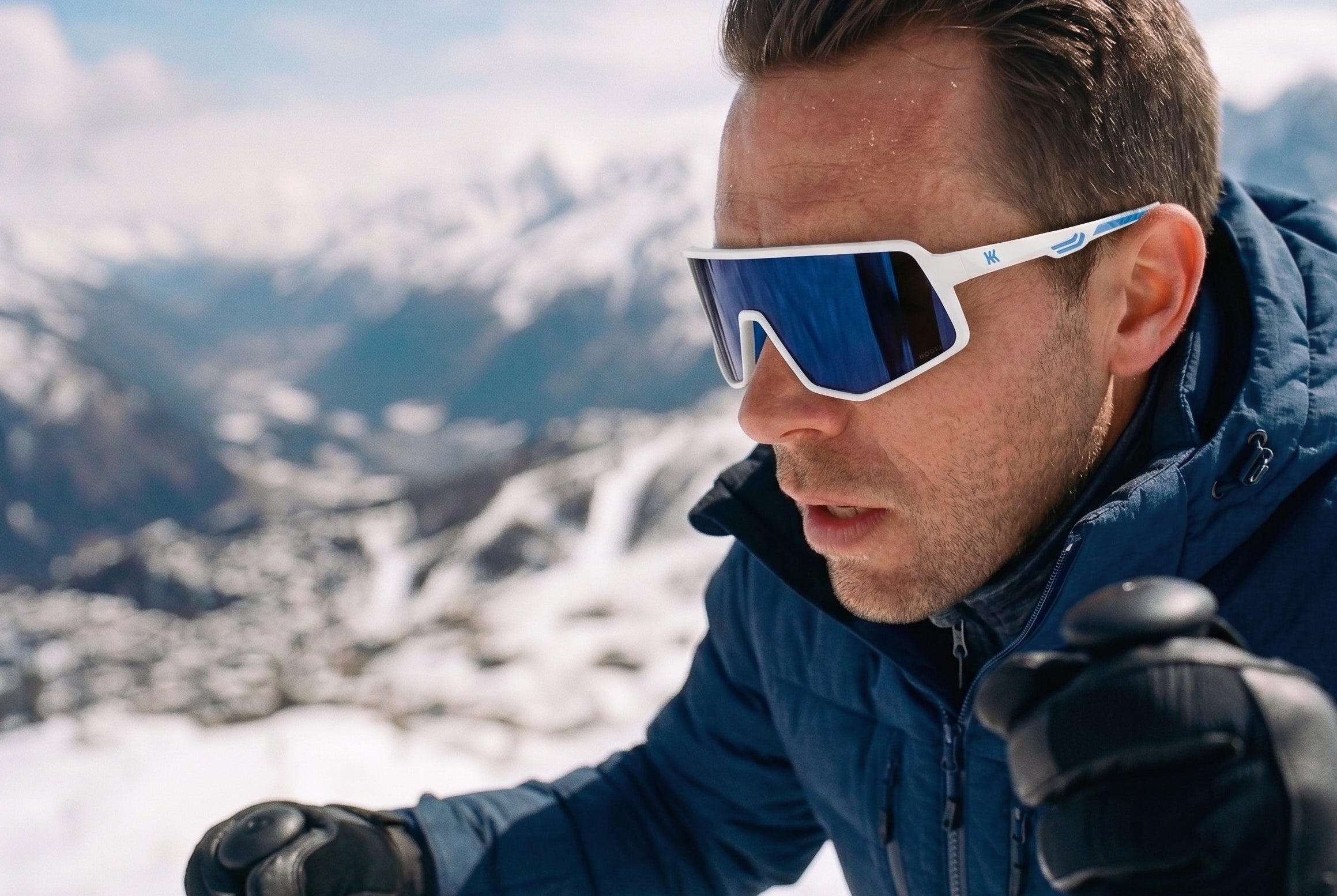 A man in a snowy mountain setting wearing white-framed ski sunglasses with blue mirrored lenses, a blue jacket, and black gloves. Film border graphics are visible on the left and right edges.