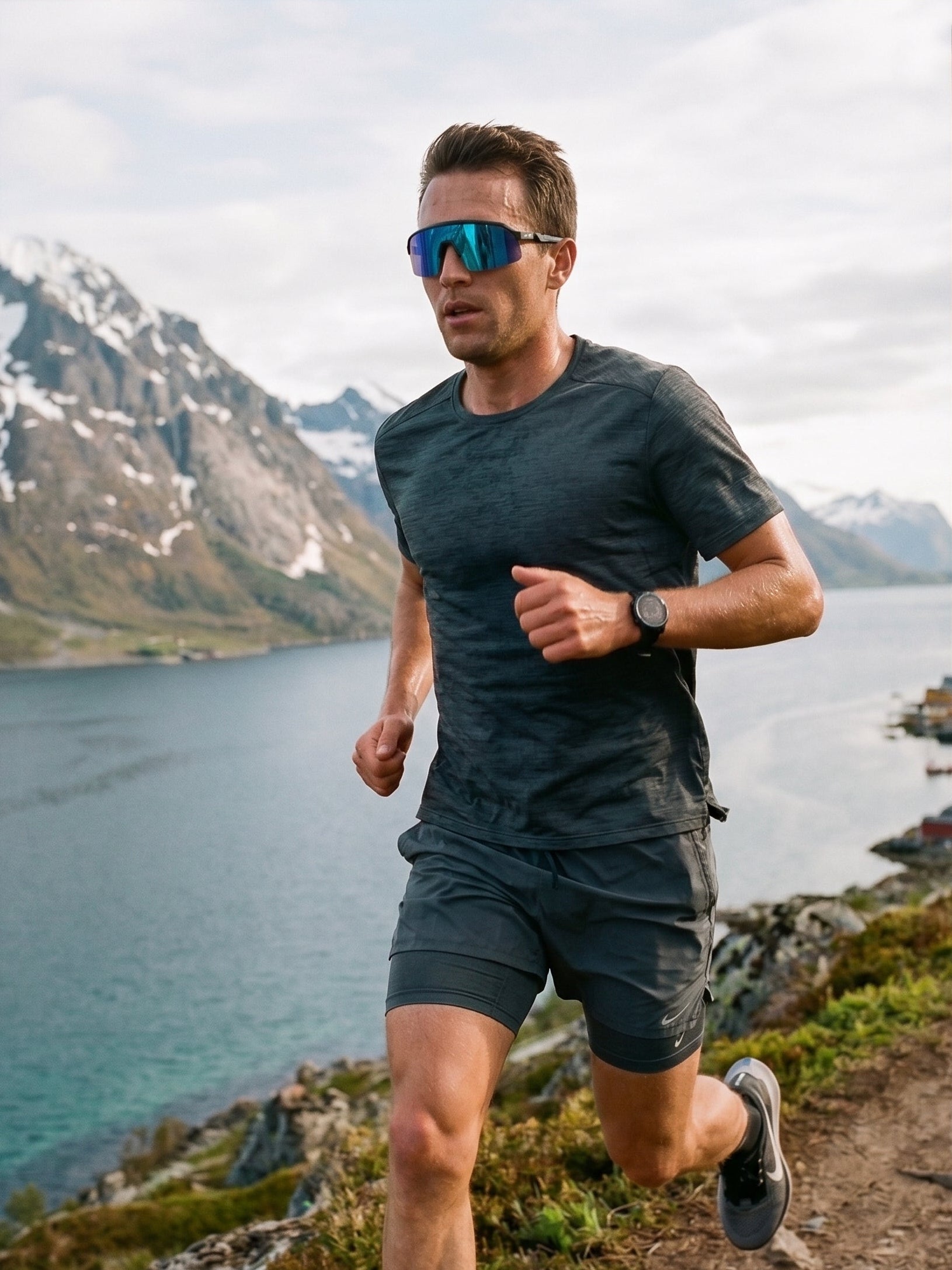 A man running outdoors near a lake and mountains, wearing a dark gray athletic t-shirt and matching shorts, with reflective Mokki sunglasses and a watch.