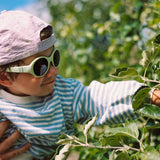 Child wearing Mokki Click & Change sunglasses and a cap, reaching out to touch leaves in an orchard.