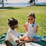 Two young girls sitting on a blanket in a park wearing Mokki sunglasses