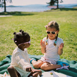 Two young girls sitting on a blanket in a park wearing Mokki sunglasses