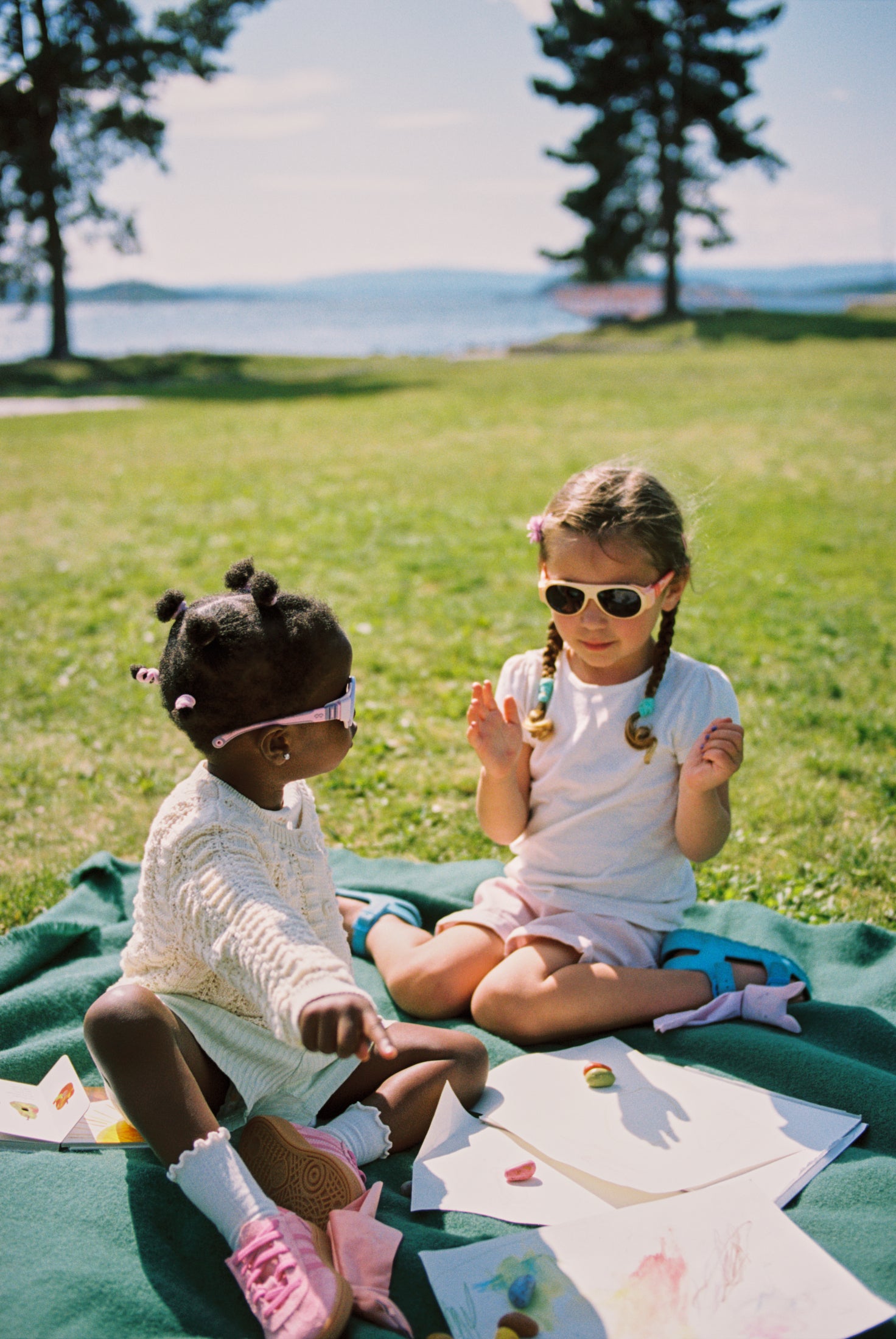 Two young girls sitting on a blanket in a park wearing Mokki sunglasses