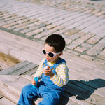 Child wearing Mokki Click & Change sunglasses and blue overalls sitting on a wooden bench.