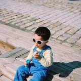 Child wearing Mokki Click & Change sunglasses and blue overalls sitting on a wooden bench.