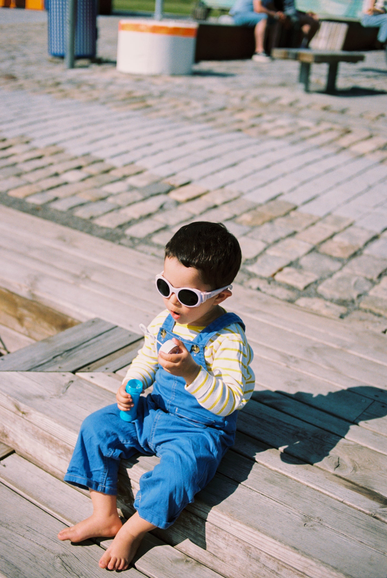 Child wearing Mokki Click & Change sunglasses and blue overalls sitting on a wooden bench.