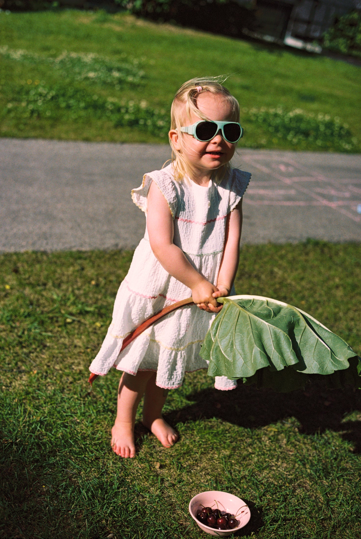 Child in a white dress holding a large leafy vegetable outdoors on grass wearing Click & Change sunglasses