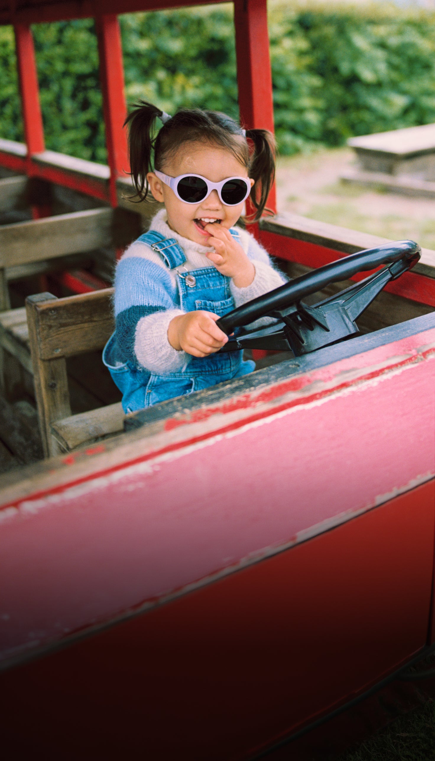 A young child with pigtails wearing round white Mokki Click & Change sunglasses and blue denim overalls sits in a red vintage-style toy bus, holding the black steering wheel.