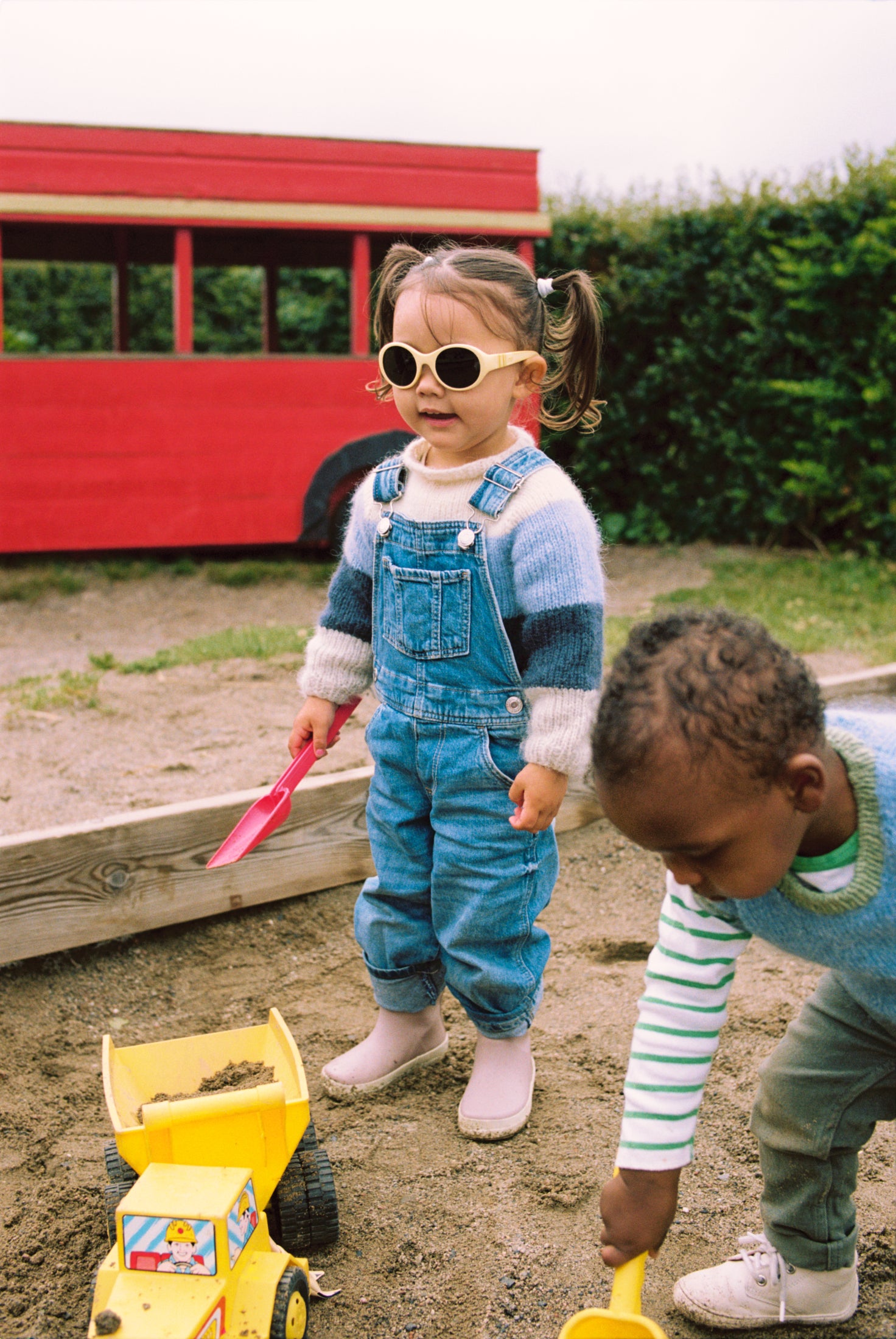 Two children wearing Mokki Click & Change sunglasses playing in a sandbox with a red train in the background
