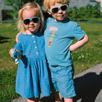Two children in blue outfits standing outdoors with Mokki Click & Change sunglasses on.