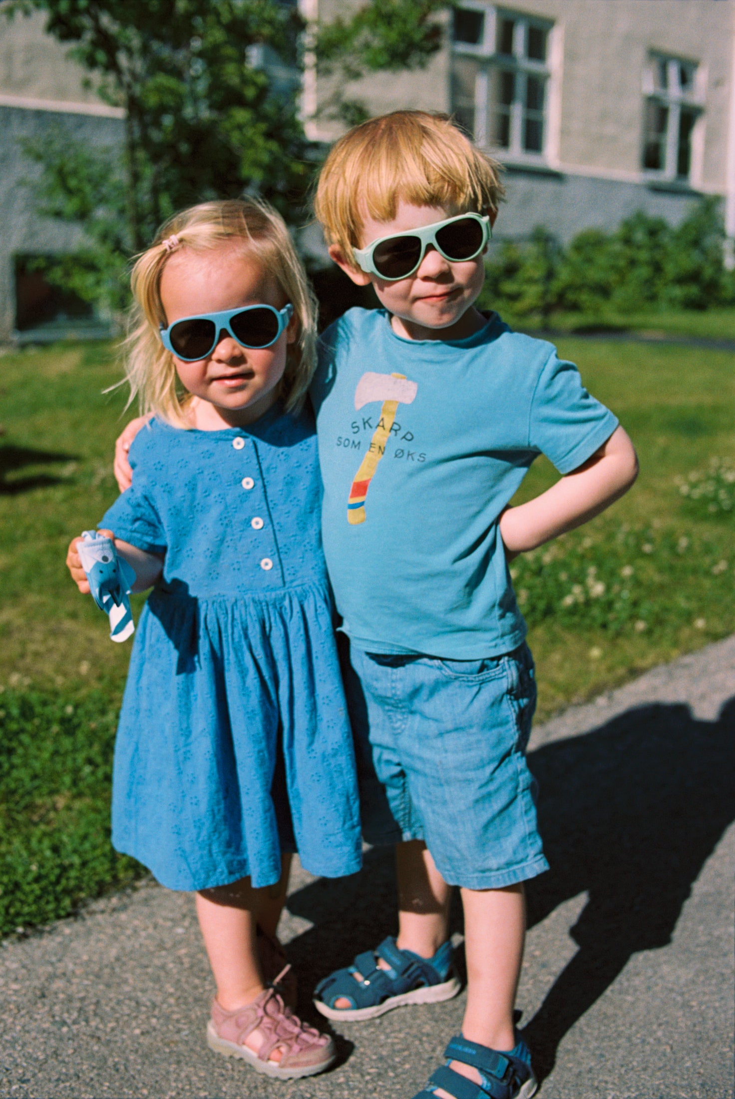 Two children in blue outfits standing outdoors with Mokki Click & Change sunglasses on.