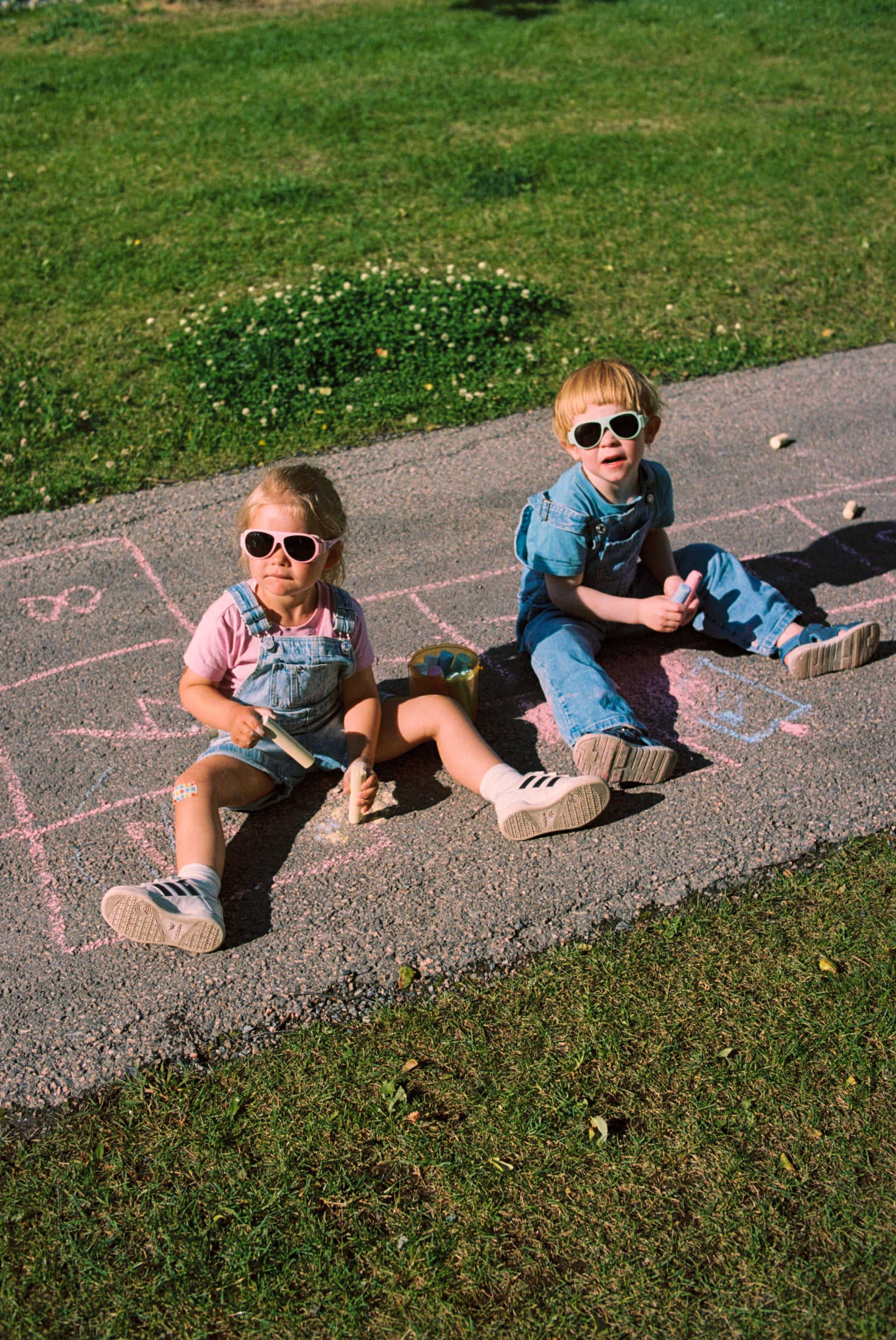Two children sitting on a sidewalk with chalk drawings, wearing Mokki sunglasses.