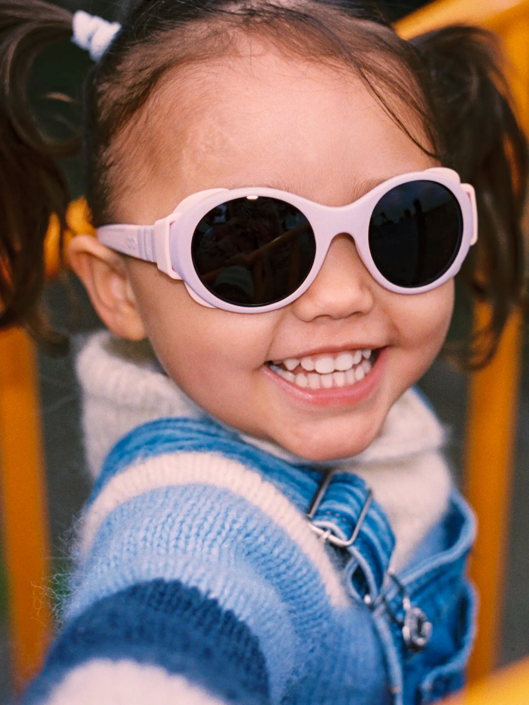 Child wearing pink Mokki sunglasses and a blue jacket on a playground.