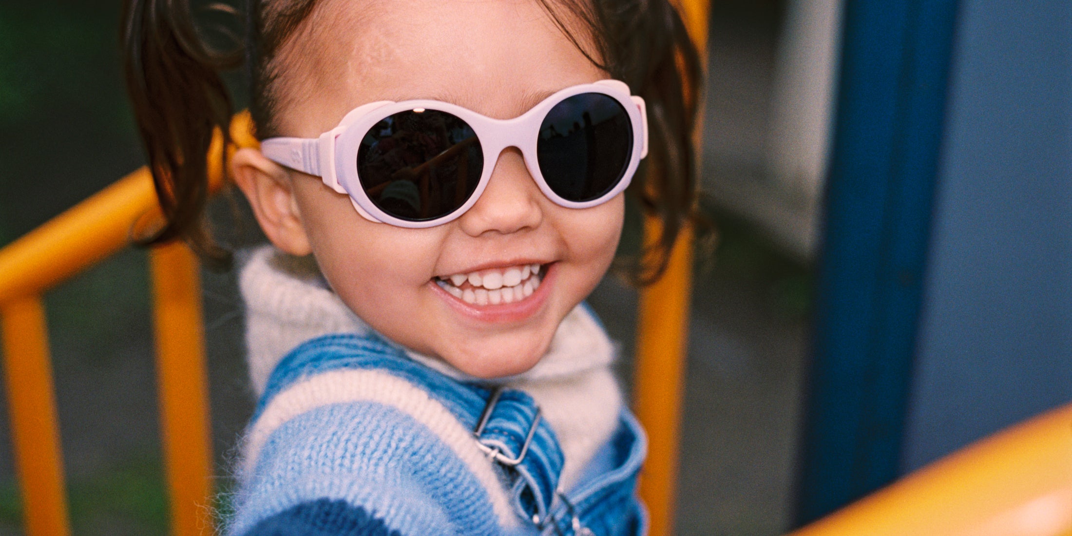 Child wearing pink Mokki sunglasses and a blue jacket on a playground.