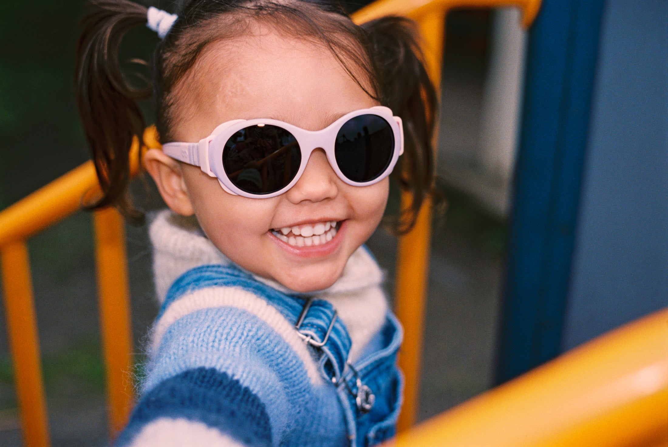 Child wearing pink Mokki sunglasses and a blue jacket on a playground.