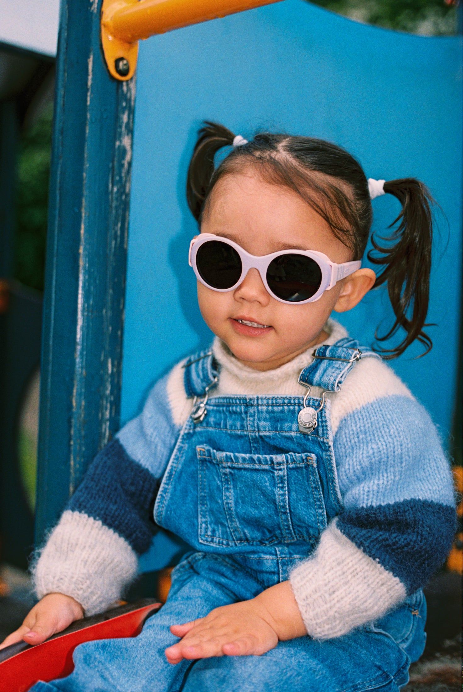 Child wearing Mokki Click & Change sunglasses and a blue outfit sitting on a playground slide.
