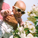 Baby wearing Mokki Click & Change sunglasses being held by an adult in a garden with white flowers.