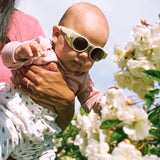 Baby wearing Mokki Click & Change sunglasses being held by an adult in a garden with white flowers.