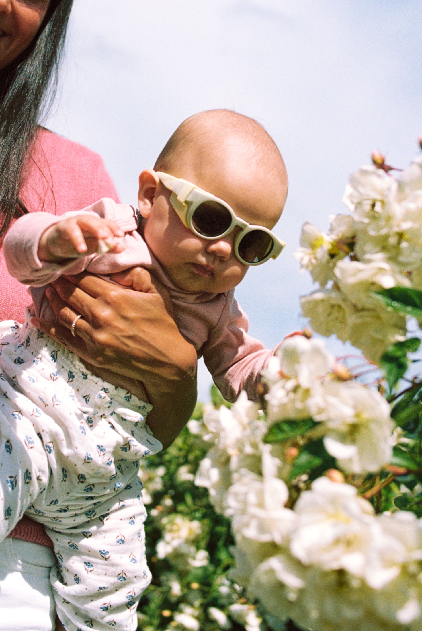 Baby wearing Mokki Click & Change sunglasses being held by an adult in a garden with white flowers.