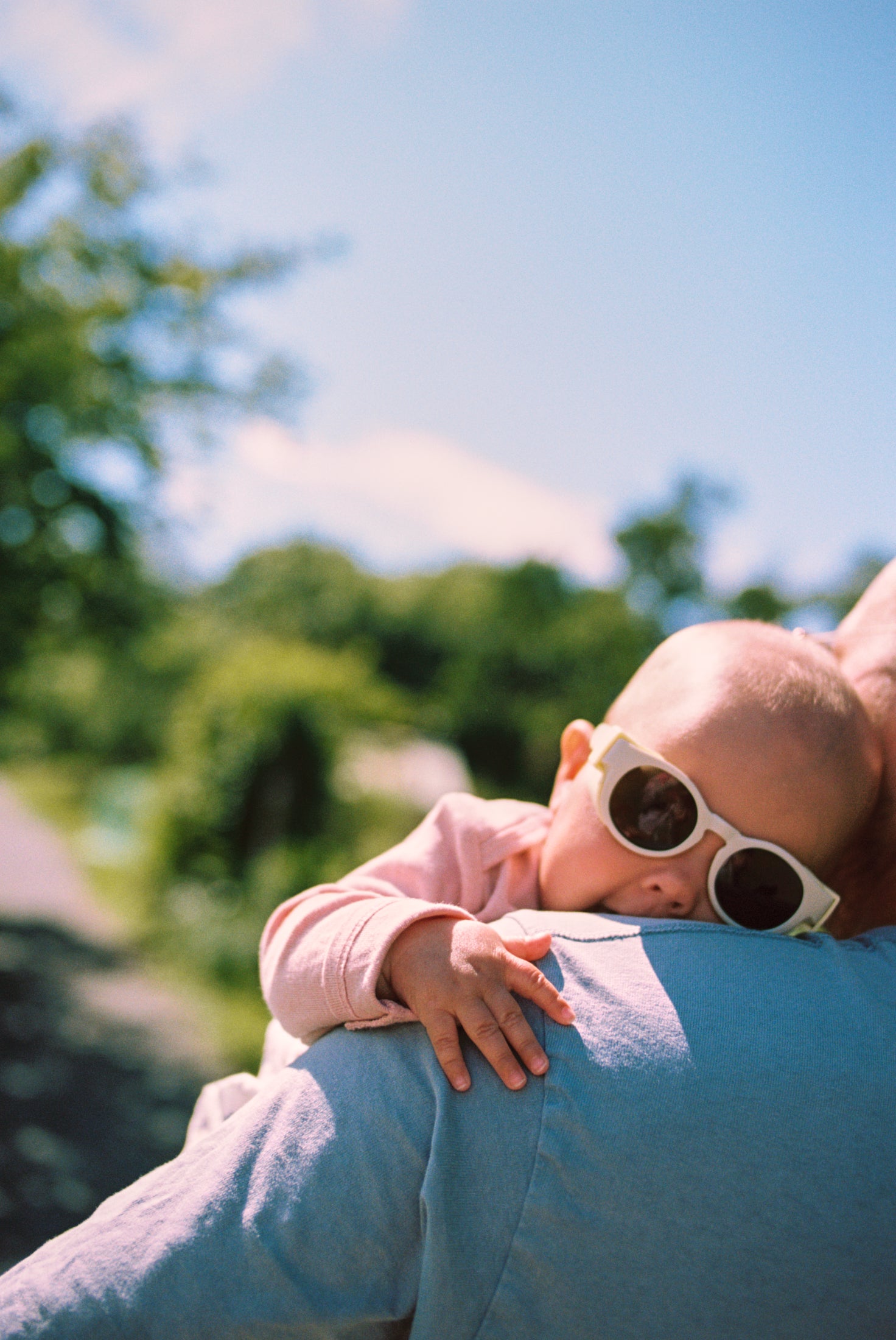 Baby wearing Mokki Click & Change sunglasses being held by an adult outdoors with trees and blue sky in the background