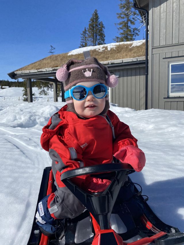 Child in a red snowsuit and Mokki Click & Change Sunglasses sitting on a snowmobile in a snowy landscape.