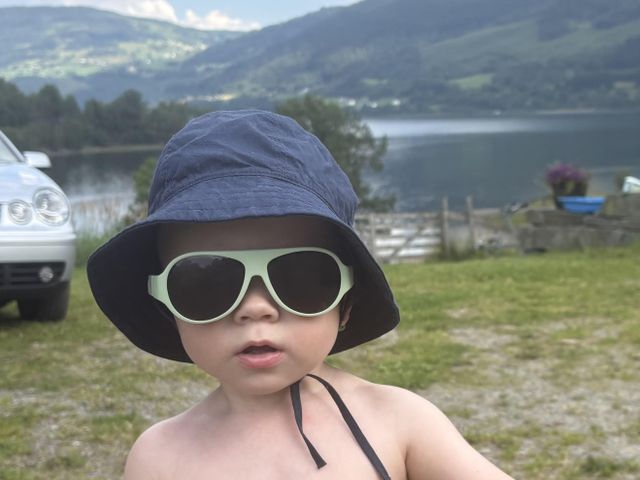 Child wearing Mokki Click & Cange sunglasses and a blue hat in a scenic outdoor setting with mountains and a lake.